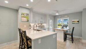 A modern kitchen with white cabinetry, a central island, and a dining area with two chairs. - The Riverside Club - B204 (Marco Island)