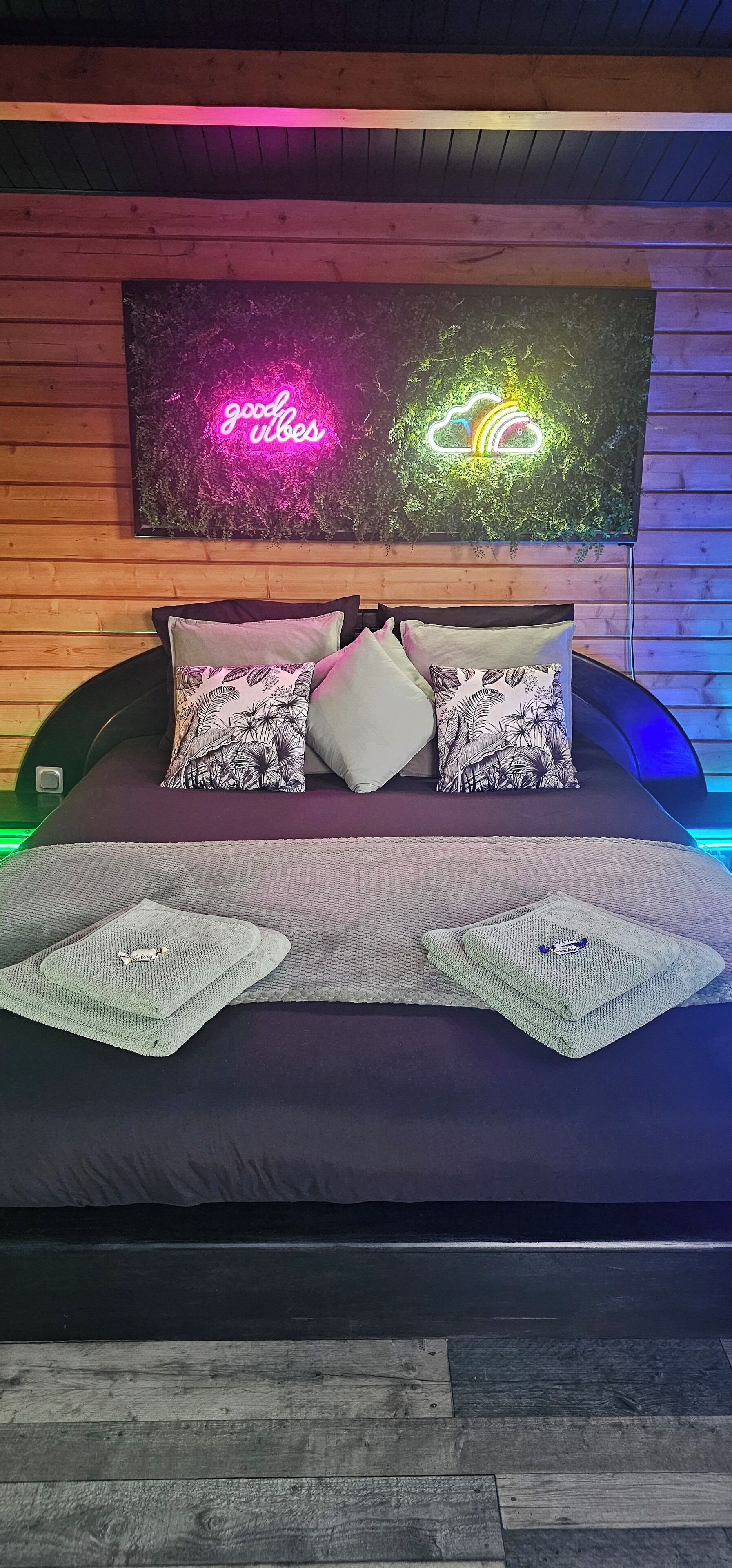 En säng med en mörk madrass, två kuddar med tropiska mönster och en stickad pläd. Ovanför sängen hänger en neonskylt med texten ”goda vibbar” och ett regnbågmoln.