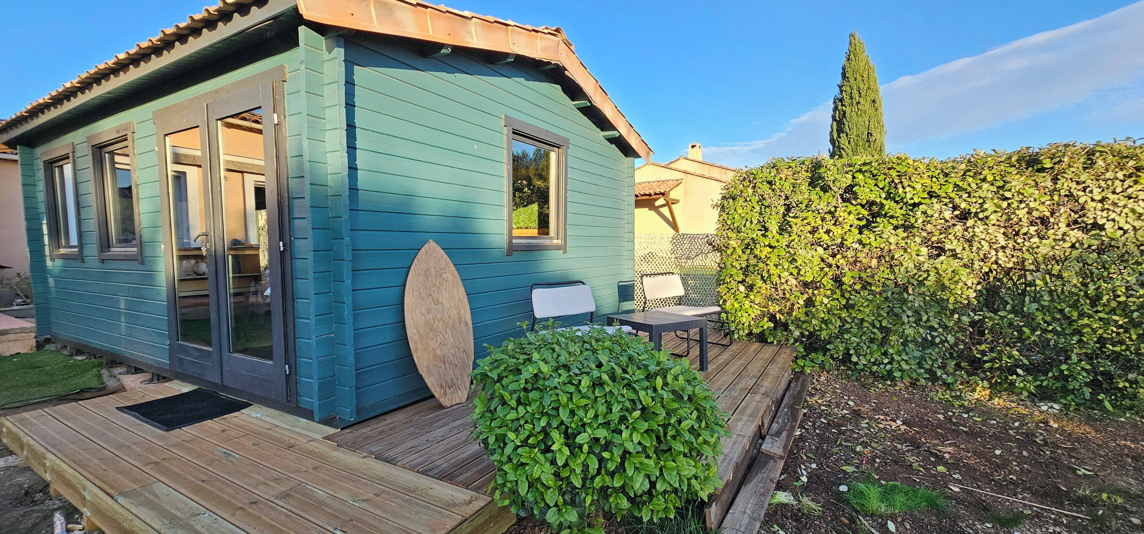 Een klein, gelijkvloers huis met een blauwe gevel, een houten terras en een tuin met een haag en een hoge boom.