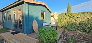 Una casa pequeña de un solo piso con fachada azul, una terraza de madera y un jardín con seto y un árbol alto.
