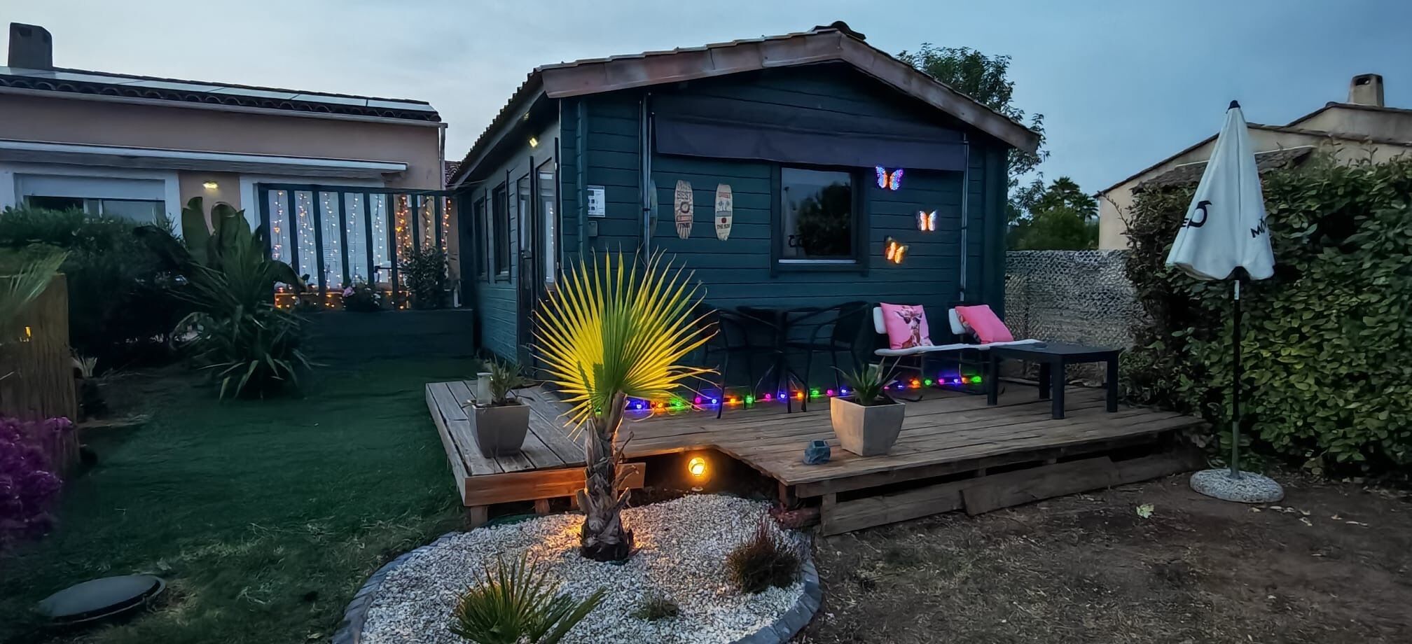 A wooden deck with seating and a small palm tree, surrounded by a garden with a house in the background.