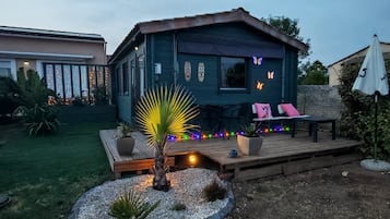 A wooden deck with seating and a small palm tree, surrounded by a garden with a house in the background.
