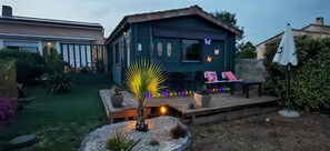 Une terrasse en bois avec des sièges et un petit palmier, entourée d’un jardin avec une maison en arrière-plan.