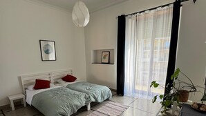 Quarto com cama, mesinha de cabeceira, luminária pendente, janela com cortinas e um vaso com planta.