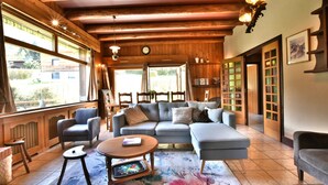 A cozy living room with a sectional sofa, a coffee table, and a rug. There are large windows with curtains, wooden beams on the ceiling, and a clock on the wall. - Authentic chalet at the foot of the Mont Blanc range (Les Houches)
