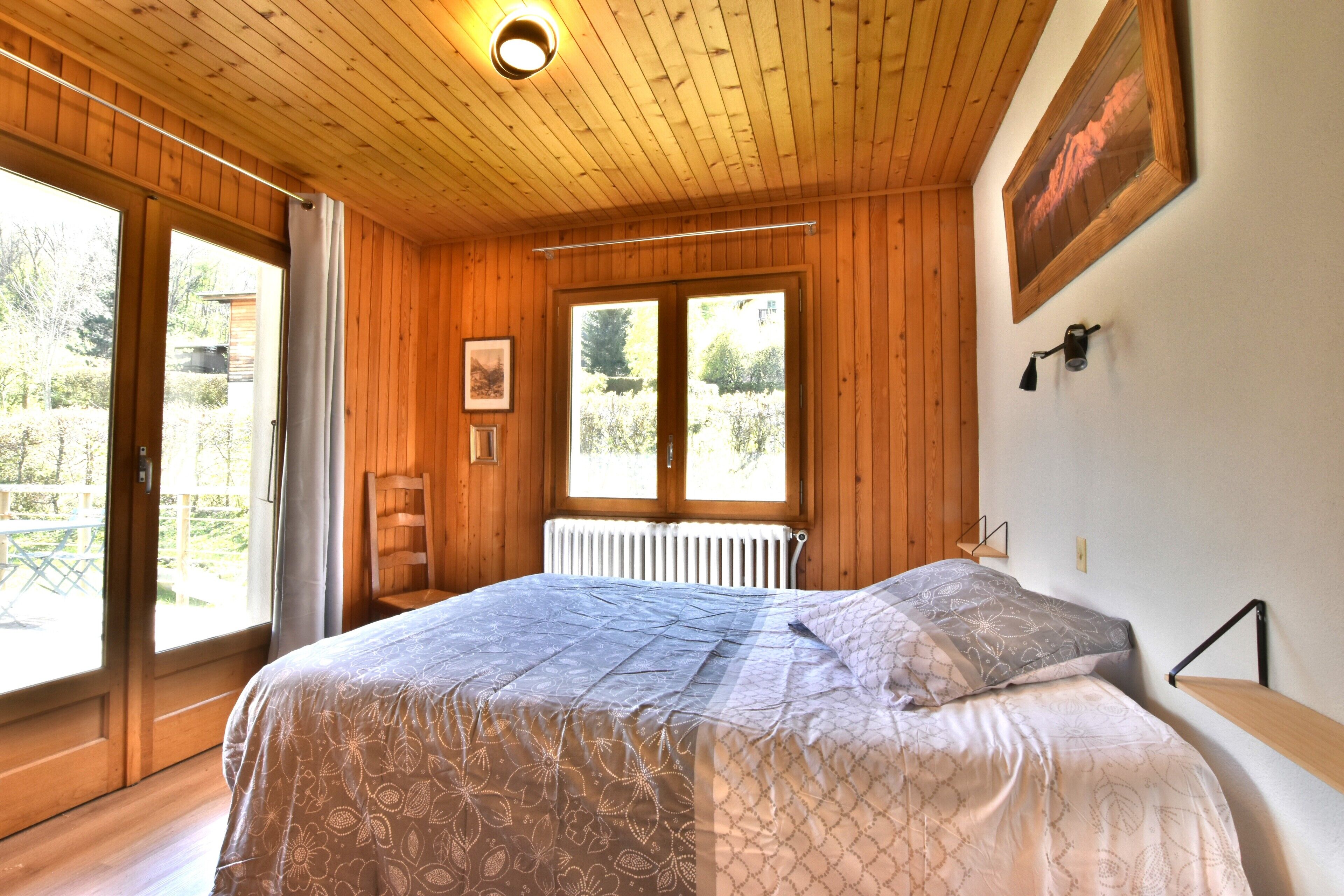 Ein Schlafzimmer mit Holzvertäfelung, einem Bett, einem Heizkörper und einem Fenster mit Blick auf Grünflächen.