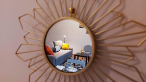 Un miroir rond, doté d’un cadre géométrique, reflète une pièce dans laquelle on aperçoit un canapé, une lampe et une petite table.