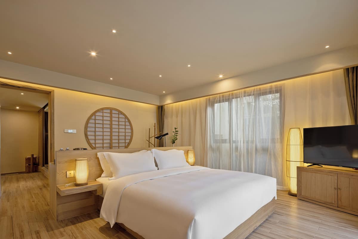 A modern hotel room with a large bed, bedside tables with lamps, a TV, and a large window with sheer curtains.