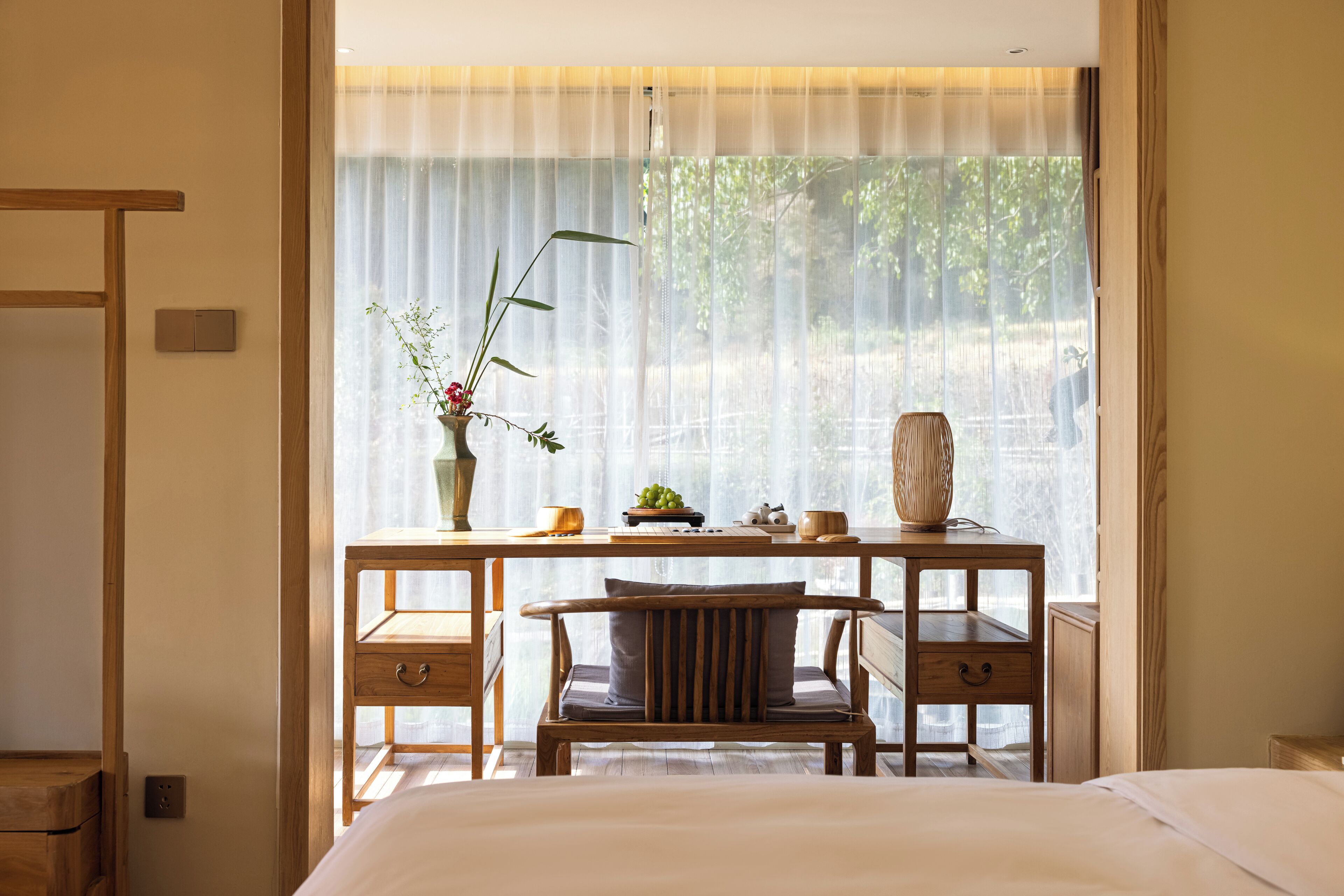 A bedroom with a bed, wooden desk, chair, and a window with sheer curtains.