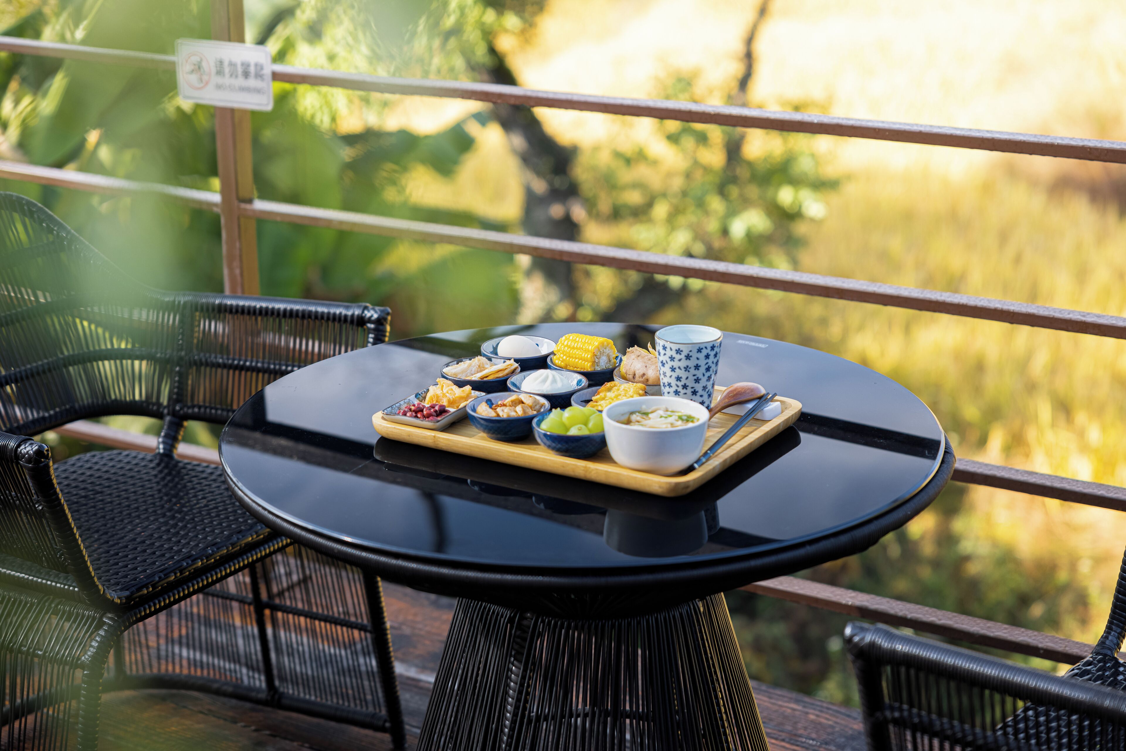 A round table with a tray of food items including corn, chips, and bowls of dips, set on a balcony with wicker chairs and a view of greenery.
