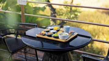 A round table with a tray of food items including corn, chips, and bowls of dips, set on a balcony with wicker chairs and a view of greenery.