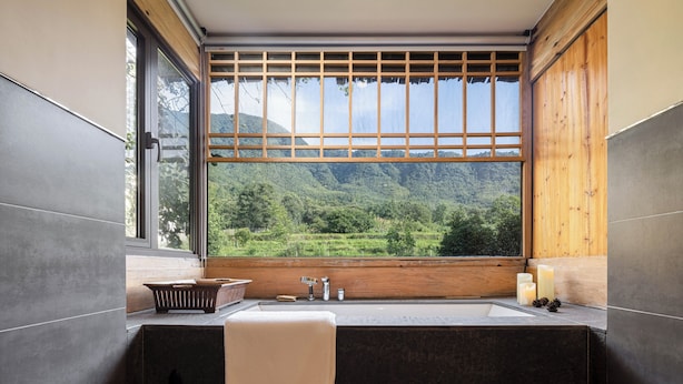 A bathroom with a bathtub, a window with a view of a mountainous landscape, and a wooden shelf with toiletries. - Tachuan Resort (Huangshan)