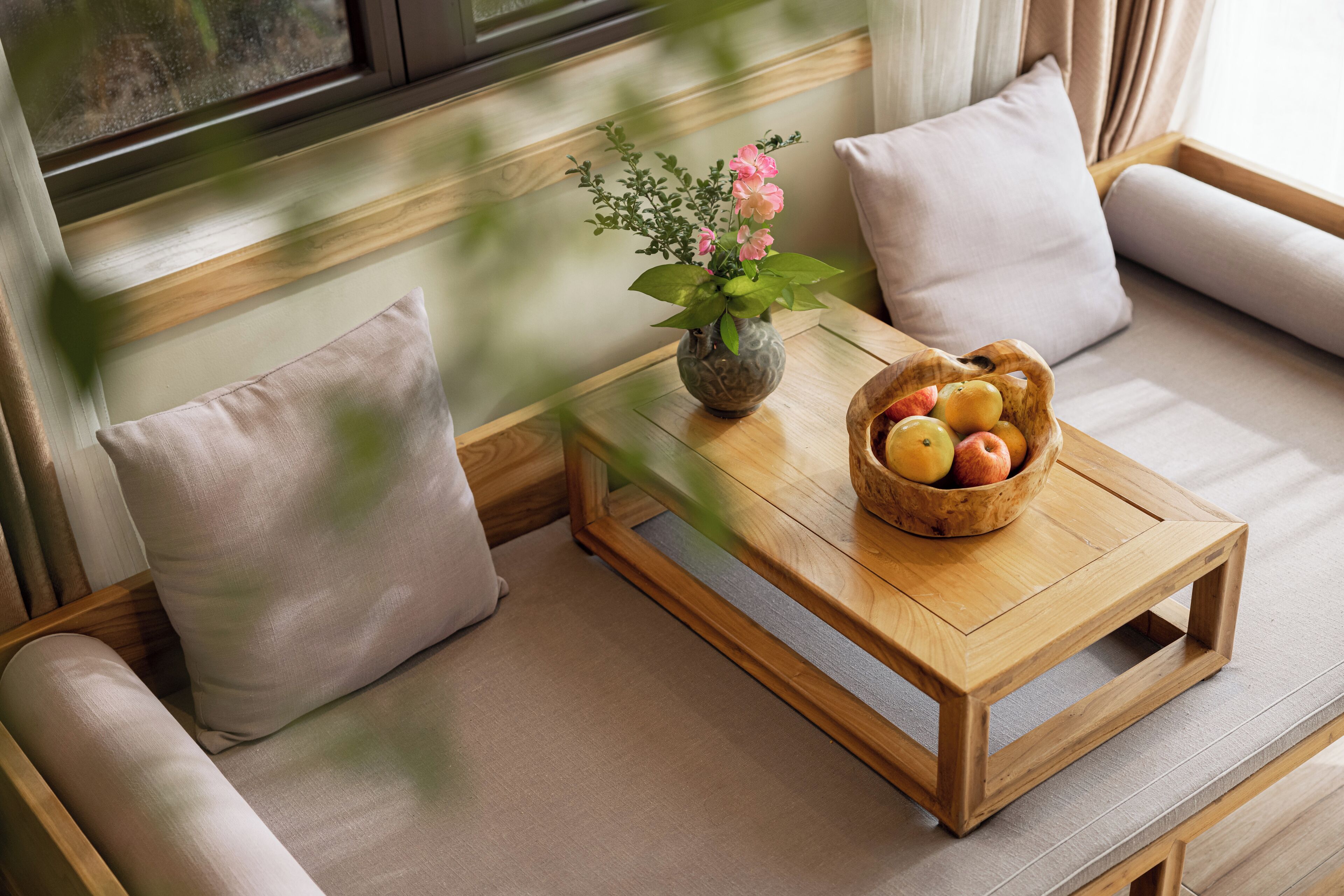 A wooden table with a vase of flowers and a bowl of fruit on a cushioned bench.