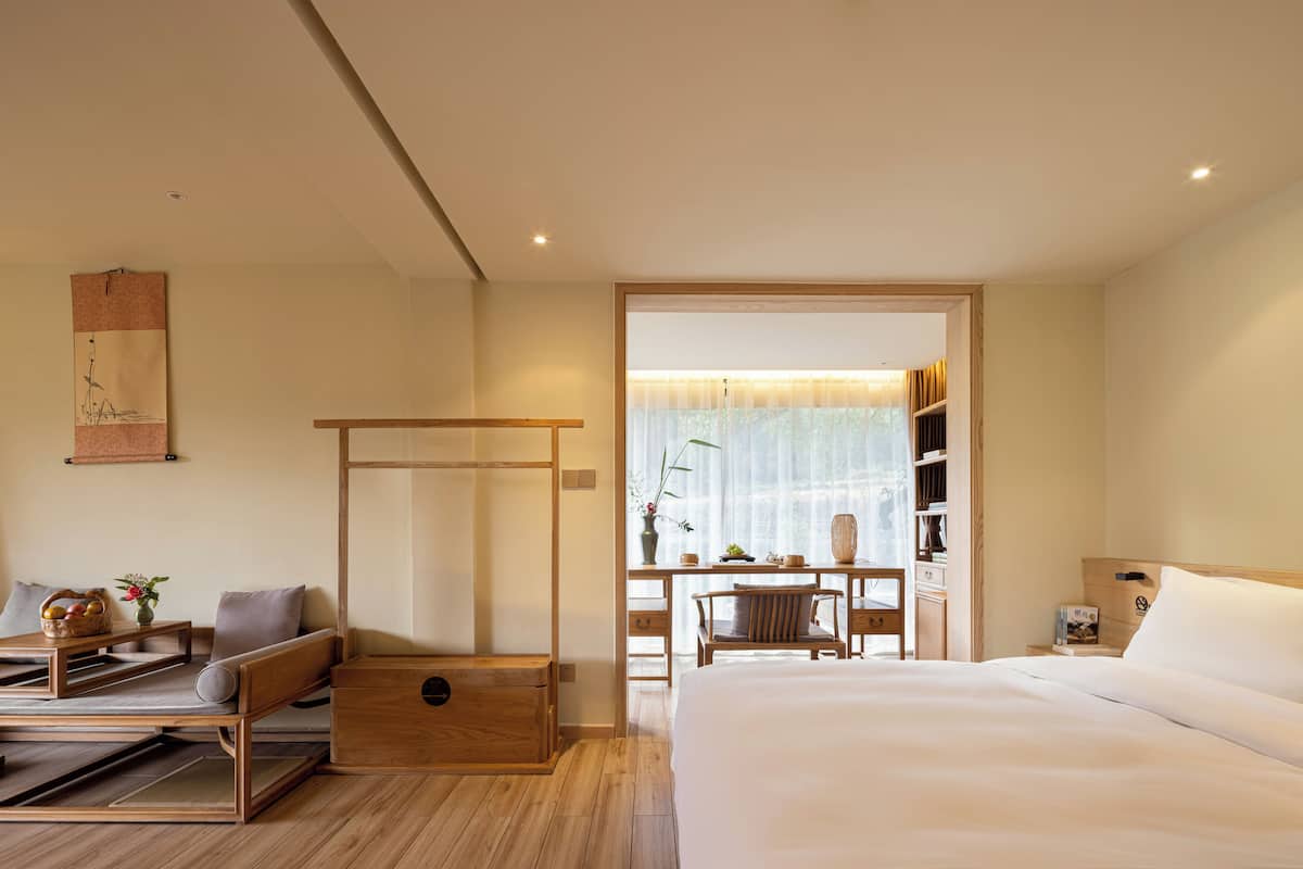 A modern hotel room with a wooden bed, a desk with a chair, a small table with a basket, and a bookshelf.