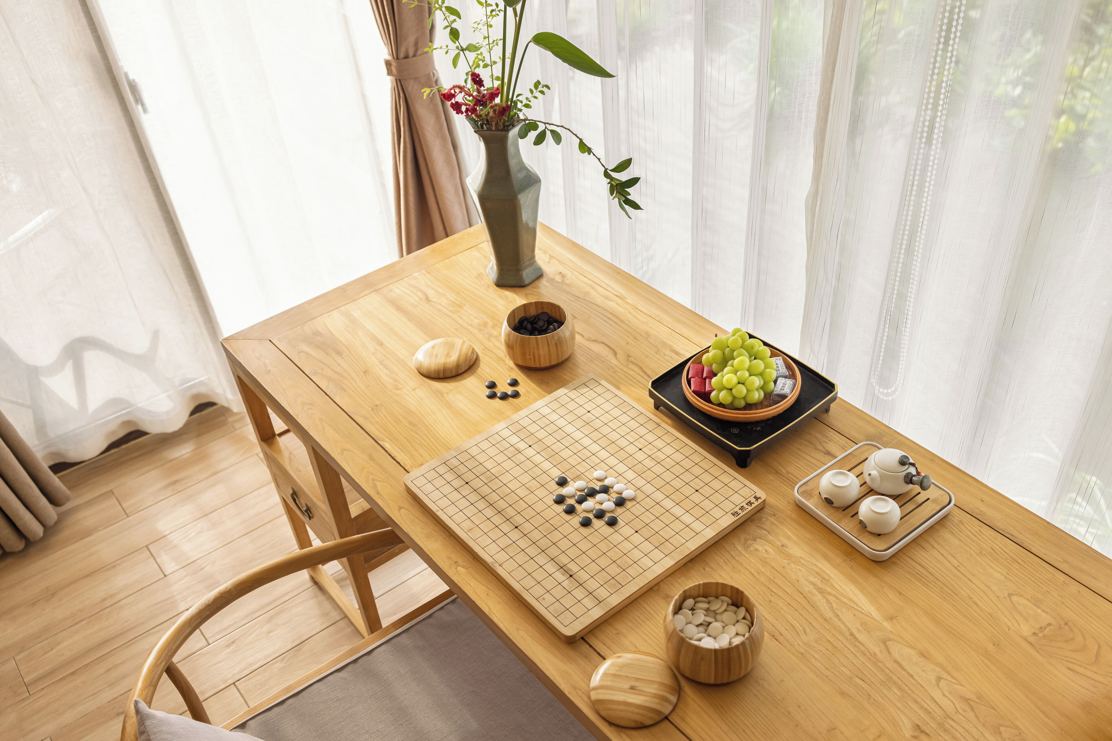 A wooden table with a Go board, a vase with flowers, a bowl of grapes, and a tray with teapots.