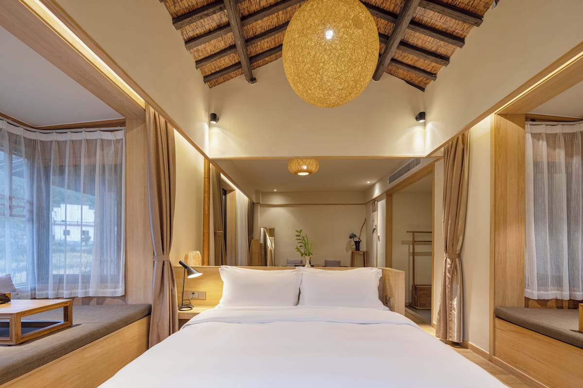 A hotel room with a large bed, wooden headboard, a ceiling light fixture, and a window with sheer curtains.