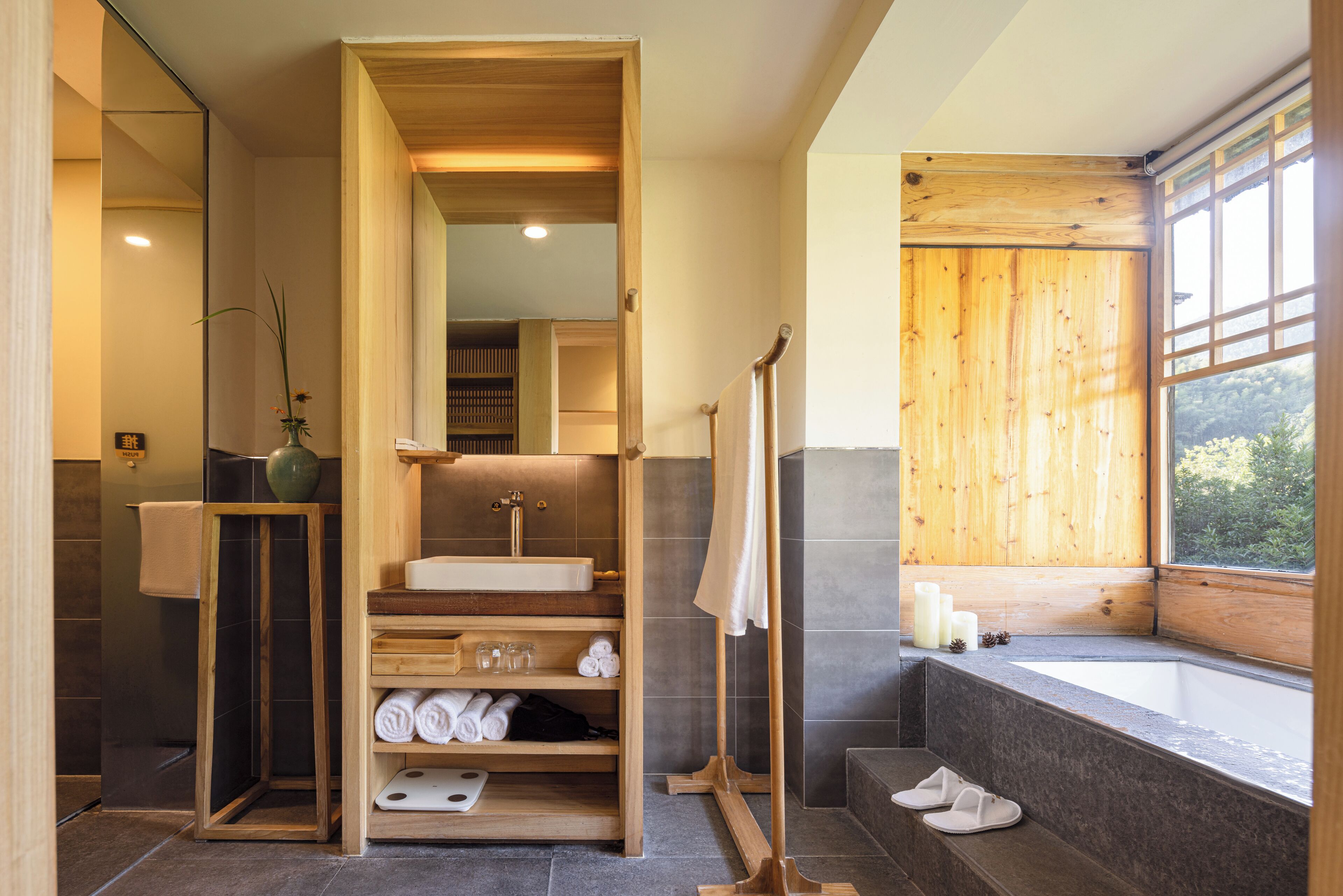 A modern bathroom with a wooden vanity, a large mirror, a bathtub, and a window with a view of greenery.