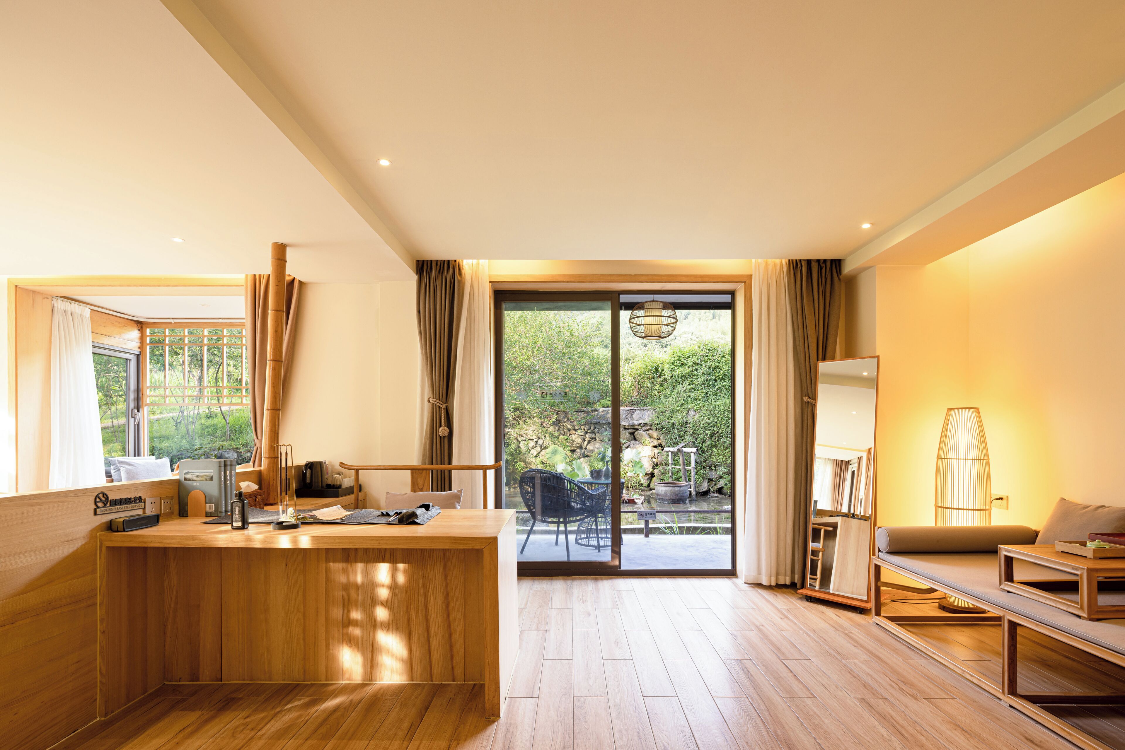 A modern hotel room with a wooden desk, a sofa, and a view of a garden through a sliding glass door.