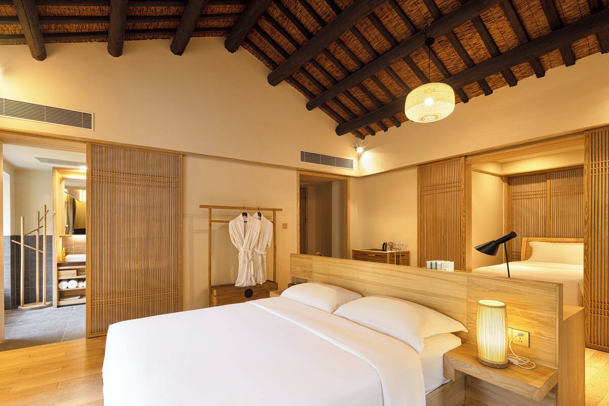 A hotel room with a large bed, wooden headboard, a bedside table with a lamp, a mirror, and a wooden wardrobe.