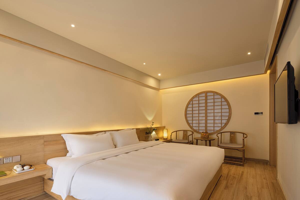 A modern hotel room with a large bed, bedside tables, a small table with a lamp, two chairs, and a wall-mounted TV.