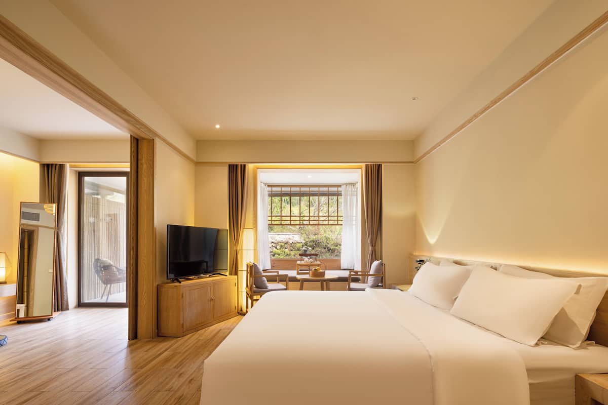 A hotel room with a large bed, a television, a desk with a chair, and a view of the outdoors through a window.