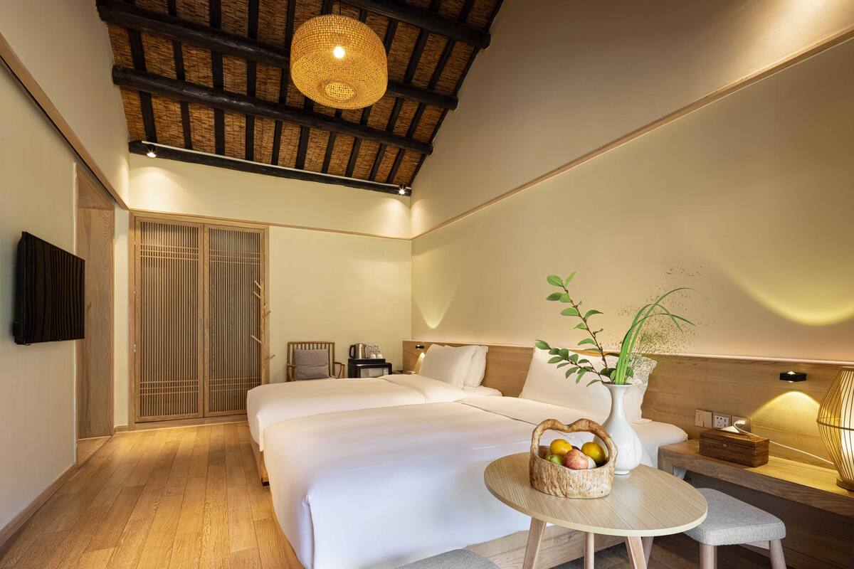 A modern hotel room with a large bed, a small round table with a fruit basket, a bedside lamp, and a wooden ceiling.