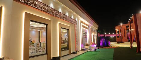 Exterior de un hotel de noche, con entradas iluminadas, un camino alfombrado de color verde e iluminación decorativa.