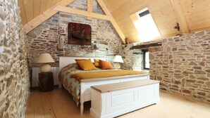 Ein Schlafzimmer mit Steinwand, Holzbalkendecke, einem Bett mit weißem Kopfteil und zwei Nachttischlampen.