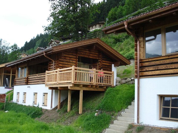 Eine Holzhütte mit einem Balkon, umgeben von viel Grün und einer Berglandschaft im Hintergrund.