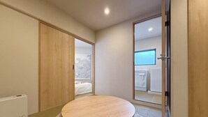Ein modernes Schlafzimmer mit einem runden Holztisch, einem Bett mit weißem Kopfteil und Blick in einen anderen Raum mit Fenster und einem weißen Gerät.