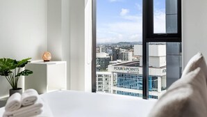 Camera d'hotel con una grande finestra che si affaccia sulla città, un letto con asciugamani ordinatamente piegati, una pianta in vaso e una pietra decorativa.