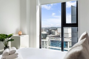 Hotellihuone, jossa on suuri ikkuna, josta avautuu näkymä kaupunkiin, siististi taitelluilla pyyhkeillä varustettu sänky, ruukussa oleva kasvi ja koristeellinen kivi.