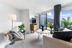Ein modernes Wohnzimmer mit einer Couch, einem Sessel, einem Couchtisch, einem Fernseher und einem großen Fenster mit Blick ins Freie.