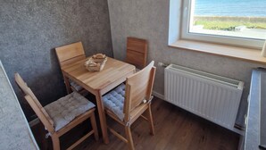 A compact kitchen with a wooden table and chairs, a clock on the wall, a window with a view of the sea, and a radiator. - Apartment 54m² for 1-4 persons directly on the dike, sea view from kitchen and living room (Dahme)