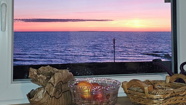 A window with a view of the sea at sunset, a glass bowl with fruits, and a wicker basket on a shelf. - Apartment 54m² for 1-4 persons directly on the dike, sea view from kitchen and living room (Dahme)