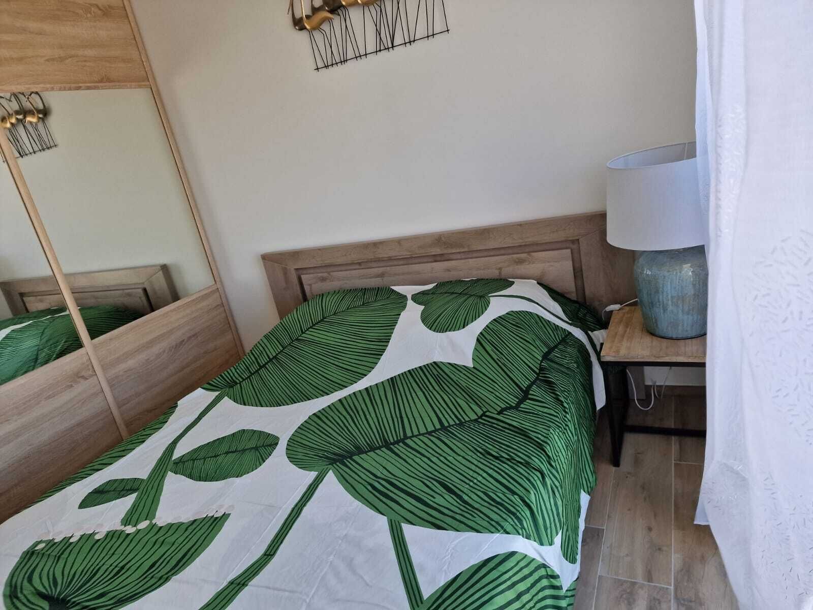 A neatly made bed with a green leaf-patterned bedspread, a wooden headboard, a bedside table with a lamp, and a mirror on the wall.