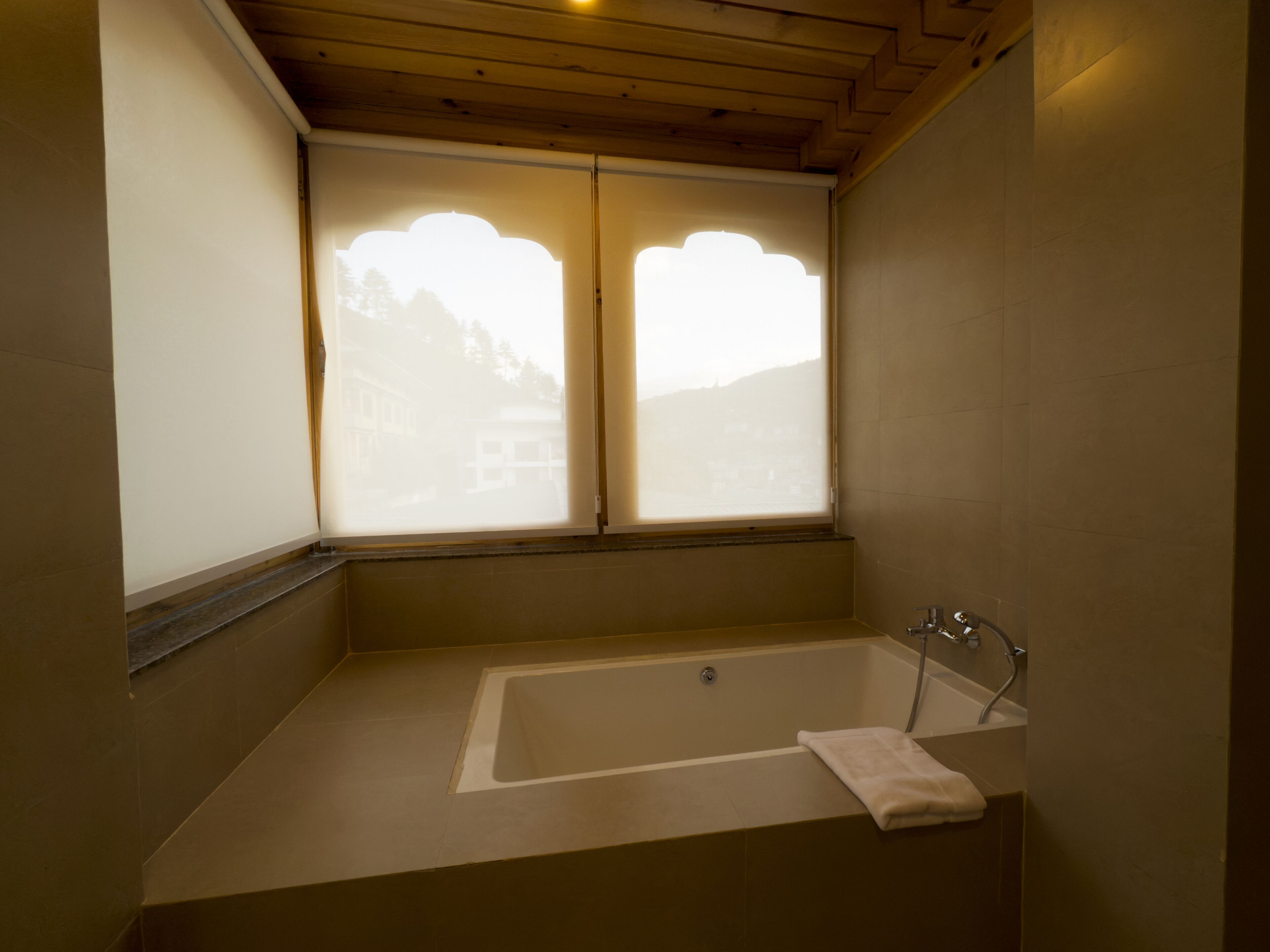 A bathroom with a bathtub, a window with a view of buildings, and wooden ceiling beams.