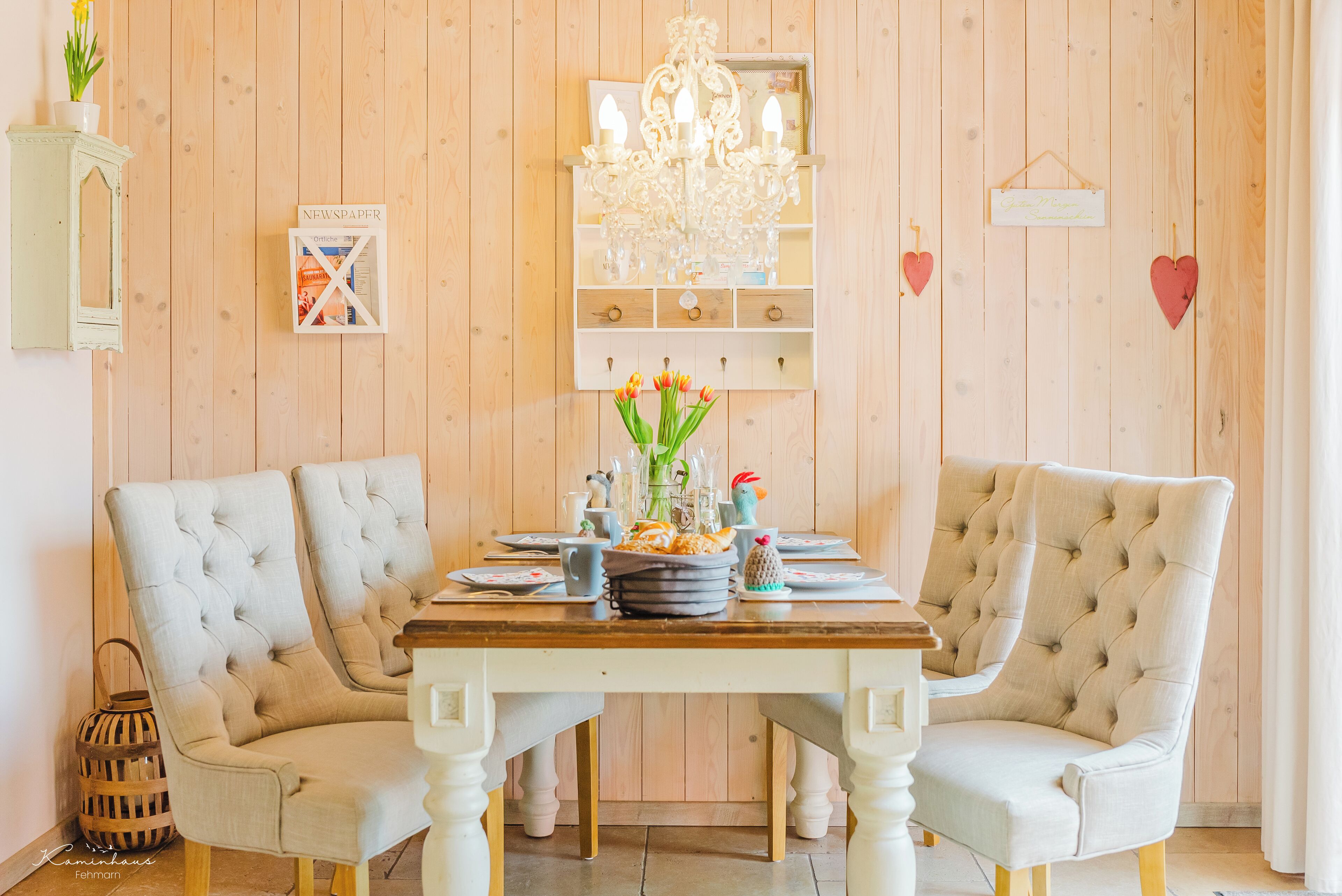 Un comedor con una mesa de madera, dos sillas tapizadas, una araña y un estante en la pared con un periódico y objetos decorativos.