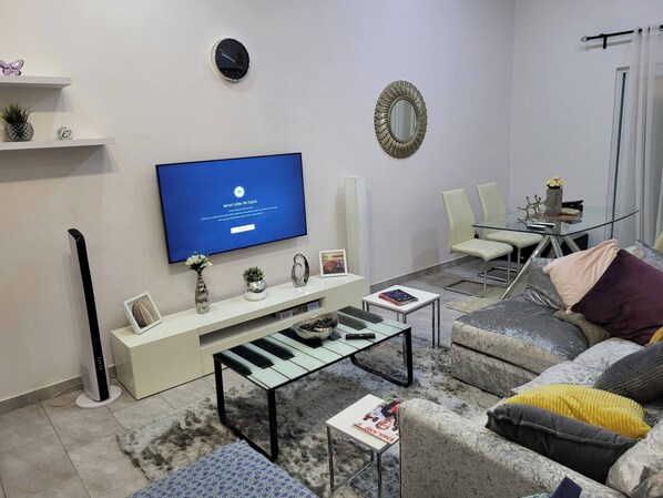 A modern living room with a flat-screen TV, a glass coffee table, a sofa, and a dining area with a glass table and chairs. - Serene Oasis in East Legon Hills - Chic 2 Bedroom Retreat (Santeo)