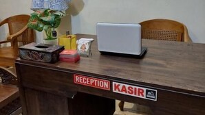 Un mostrador de recepción con la etiqueta "RECEPTION KASIR", una computadora portátil, una planta en maceta y una caja decorativa.