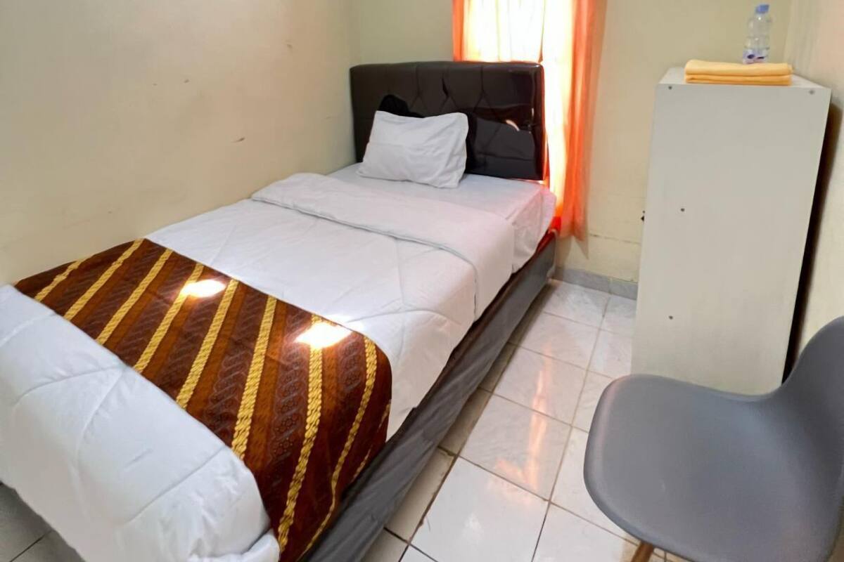 Un lit simple avec une couvre-lit à rayures, une chaise grise et une armoire blanche dans une pièce aux murs unis et au sol carrelé.