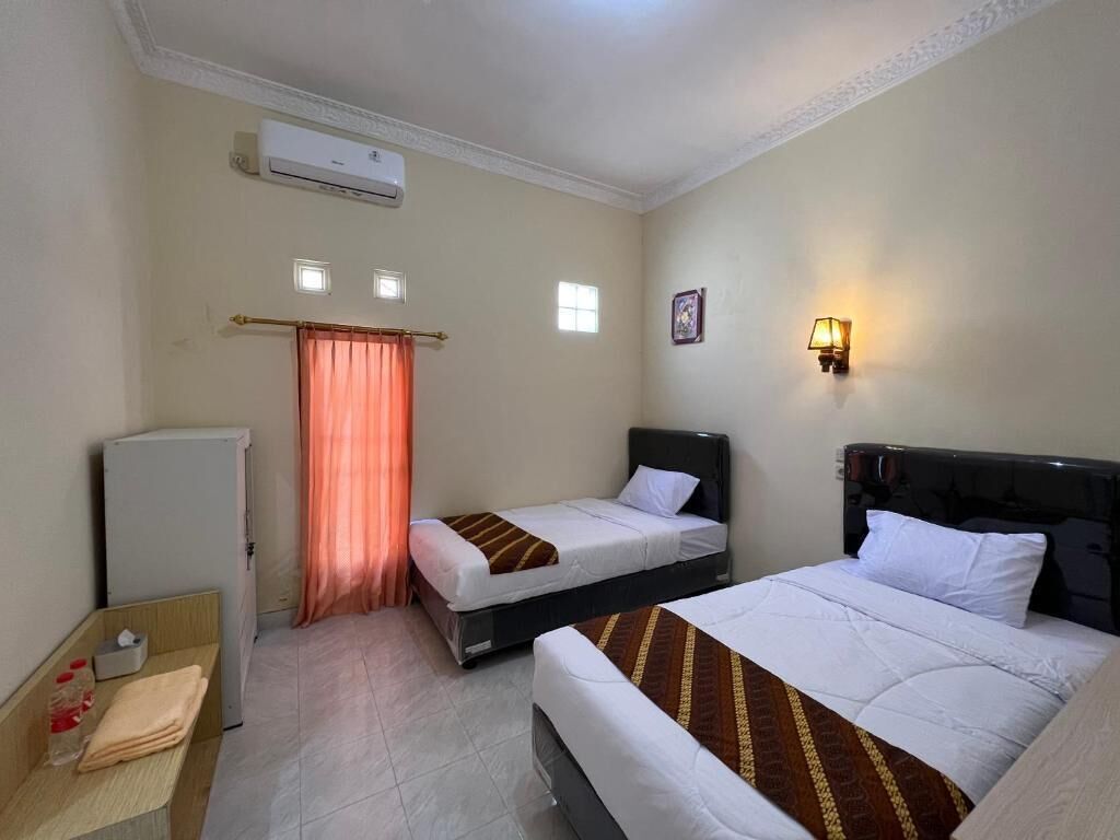 A hotel room with two beds, a small refrigerator, a bedside table, and a wall-mounted lamp.
