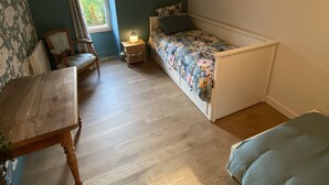 Une petite pièce agréable et chaleureuse, avec un plancher en bois, un lit recouvert d’une literie à motifs floraux, une chaise, une petite table et une fenêtre avec des rideaux.