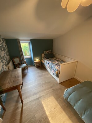 Una habitación pequeña y acogedora con piso de madera, una cama con ropa de cama floral, una silla, una mesita y una ventana con cortinas.