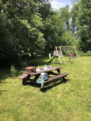 Un montaje para picnic con una mesa, cesto de picnic y un columpio en un espacio cubierto de césped con árboles.
