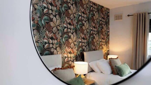 A bedroom with a floral wallpaper, a bed with pillows, a nightstand with a lamp, and a round mirror on the wall. - Vacant Nests | The Carrick  | Beach | Golf Course (Hartlepool)