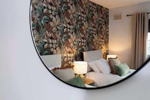 A bedroom with a floral wallpaper, a bed with pillows, a nightstand with a lamp, and a round mirror on the wall.