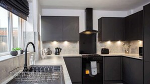 A modern kitchen with dark cabinetry, a stainless steel sink, a built-in oven, and a window with blinds. - Vacant Nests | The Carrick  | Beach | Golf Course (Hartlepool)