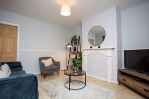 Ein Wohnzimmer mit Sofa, Sessel, rundem Tisch, TV-Schrank, Kamin und Spiegel.