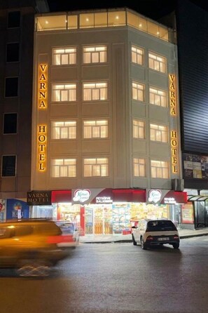 A multi-story hotel with illuminated signage, a ground-level retail area, and a parking lot. - Varna Hotel (Kesan)